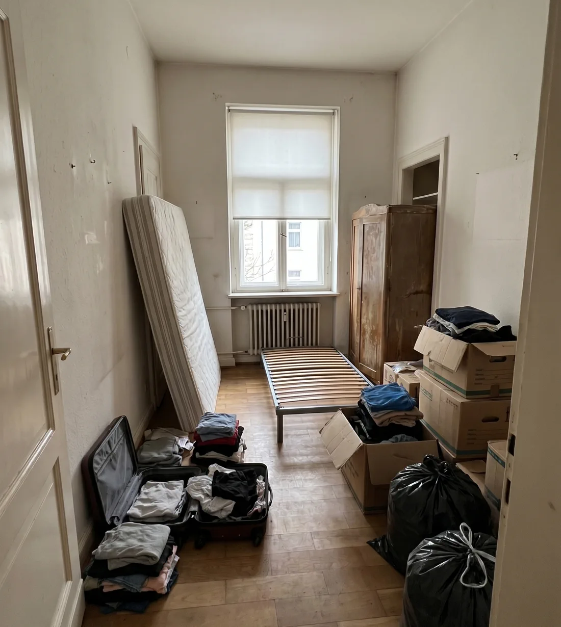 Beispieldarstellung: Altbau-Schlafzimmer vor der Räumung – Matratze, Kleiderschrank, Koffer und Umzugskartons auf Parkett.