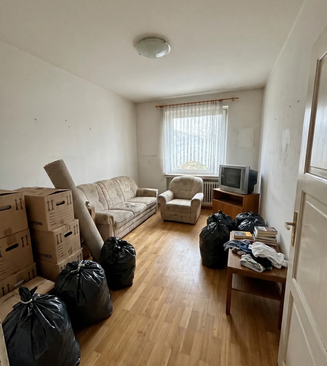 Beispieldarstellung: Wohnzimmer vor der Haushaltsauflösung – Sofa, Sessel, Umzugskartons und Müllsäcke auf Parkett.